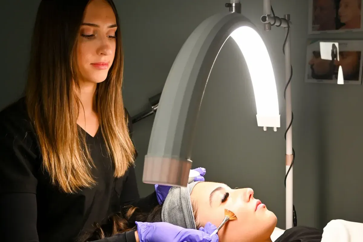 An aesthetician painting a peel onto a client’s cheek with a fan brush.