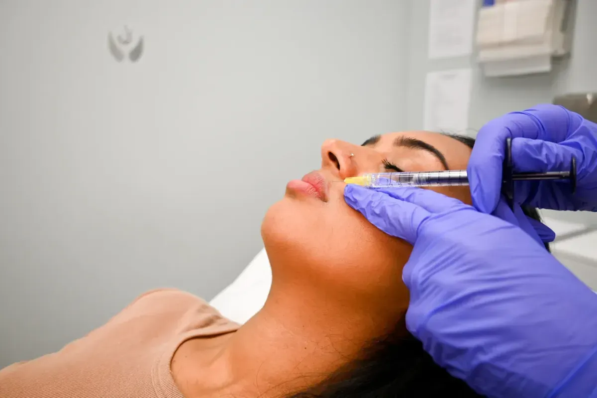 Gloved hands placing filler beside a client’s nose and lip, side view.